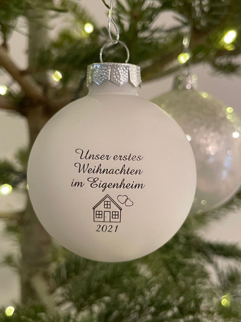May include: A matte white spherical Christmas bauble with a silver cap. The front features black printed text that reads "Unser erstes Weihnachten im Eigenheim" above a line drawing of a house with two small hearts and the year 2021 below.