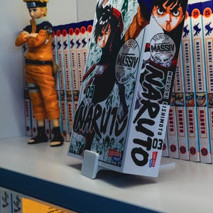 Könnte beinhalten: Ein Regal mit einer Sammlung von Manga-Comics, darunter ein großer Band von Naruto, Band 3. Der Comic ist auf Deutsch und trägt den Text "MASSIV" auf dem Cover. Eine kleine Naruto-Figur steht auf dem Regal.