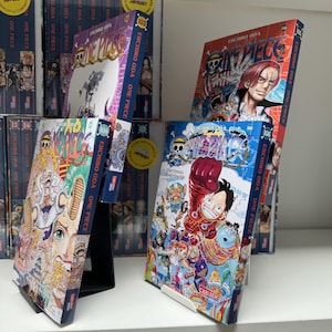 Könnte beinhalten: Eine Sammlung von One Piece Manga-Bänden, mit den Covern, die verschiedene Charaktere aus der Serie zeigen. Die Bände sind in einem Stapel angeordnet, wobei der oberste Band Luffy, den Hauptcharakter, in einem rot-weißen Outfit zeigt. Die Bände sind alle auf Japanisch.