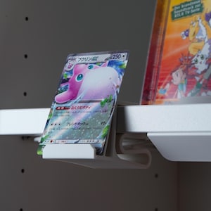 Könnte beinhalten: Ein weißer Plastik-Kartenhalter mit einer rosa und blauen Pokemon-Karte mit dem Text "プクリン 250*" und "ふくらむボディ".