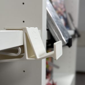 May include: White plastic shelf bracket with a hook, designed to hold items on a shelf. The bracket has a small, raised symbol of a drop of water.