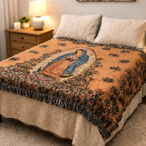 May include: A woven throw blanket featuring an image of Our Lady of Guadalupe in the center, surrounded by floral patterns in shades of orange, blue, and green, with a fringed edge, draped over a bed with two white quilted pillows.