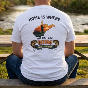 Può includere: T-shirt bianca con una grafica raffigurante lo stato della West Virginia, una scena di pesca e la scritta "HOME IS WHERE THE FISH ARE BITING WEST VIRGINIA". La maglietta è indossata da una persona seduta su un tavolo da picnic in legno.