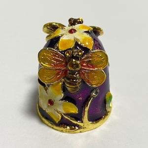 Puede incluir: Un dedal decorativo con base morada, adornado con una mariposa dorada y flores amarillas. El dedal tiene ribetes dorados y está adornado con pequeños cristales. Un artículo de colección único.