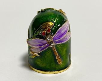 May include: A green metal thimble with a gold rim and a purple dragonfly design. The dragonfly has a clear rhinestone body and red accents.