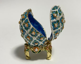 May include: An open, ornate egg-shaped trinket box. The box is blue with silver filigree, orange and green floral accents, and a gold-colored base. The interior is a deep blue.