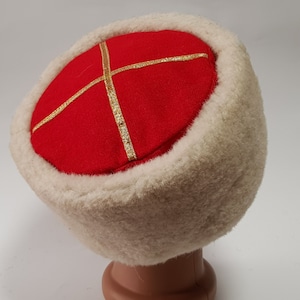 May include: A red felt hat with a gold trim and a white fur brim. The hat is on a display stand.