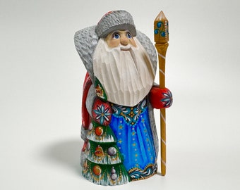 May include: Hand-carved wooden figurine of a traditional Russian Santa Claus, or Ded Moroz, wearing a red and blue robe, a white beard, and a grey fur hat. He is holding a staff with a gold top and is standing in front of a green Christmas tree.