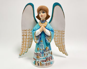 Op de afbeelding: Handgeschilderd houten engelenbeeldje met een serene uitdrukking. De engel draagt een blauwe jurk met gouden accenten en een gedetailleerd stadsgezichtontwerp. De vleugels zijn wit met gouden details en turquoise randen.