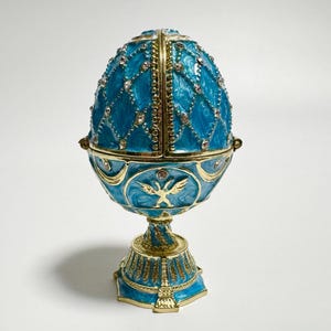 以下が含まれることがあります： ターコイズとゴールドの色合いの、装飾的な卵型の小物入れ。卵は、埋め込まれたクリスタルと金色のトリムが付いたダイヤモンドパターンが特徴です。ベースには、金の白鳥のエンブレムが付いた詳細なデザインがあります。ヒンジ付きの蓋が開きます。