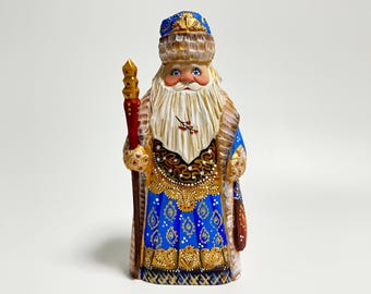 May include: Hand-carved wooden figurine of a Santa Claus wearing a blue and gold robe with a white fur trim. He is holding a staff with a red tip and has a white beard and mustache.