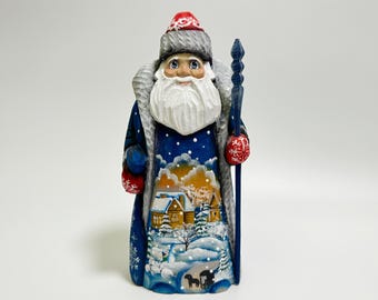 May include: Hand-carved wooden figurine of a Santa Claus wearing a blue coat with a white fur trim, a red hat, and holding a staff. The background of the figurine is a winter scene with snow-covered houses and trees.