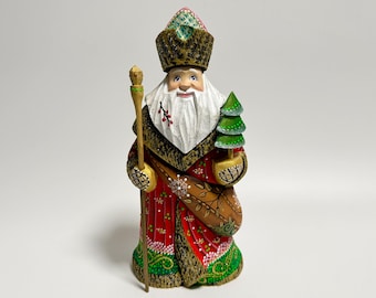 May include: A hand-carved wooden figure of a Santa Claus-like character. He wears a red coat with gold trim, a fur hat, and holds a staff and a small green Christmas tree. The figure has a white beard and a smiling expression.