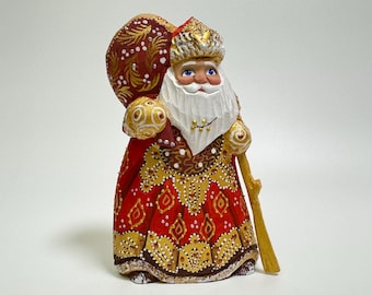 May include: A wooden figurine of a Santa Claus figure with a white beard and a red and gold robe. He is holding a staff and a bag of gifts.