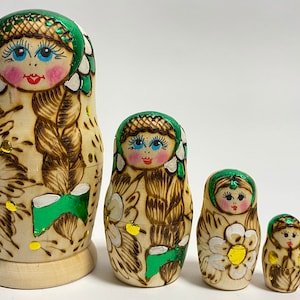 Può includere: Set di cinque bambole russe tradizionali a incastro, o bambole matrioska. Le bambole sono dipinte con motivi floreali e presentano uno schema di colori verde e oro. La bambola più grande è alta circa 15 cm.