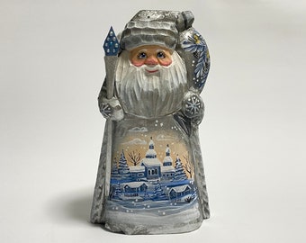 May include: A hand-carved wooden Santa Claus figurine with a white beard and a silver painted finish. He is holding a staff and a bag of gifts. The background is a winter scene with a village and snow.