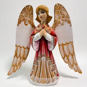 May include: A hand-painted wooden angel figurine with outstretched wings. The angel wears a red and gold dress with white sleeves and holds its hands together in prayer. The wings are white with gold and brown decorative patterns.