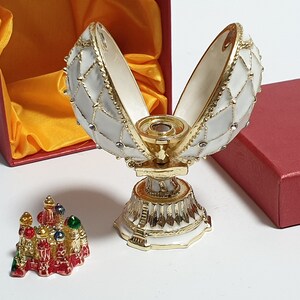 May include: A white and gold Faberg&eacute; egg with a red and gold miniature building inside. The egg is open and the building is visible. The egg is on a white stand with gold trim. The egg is in a red box with a yellow satin lining.