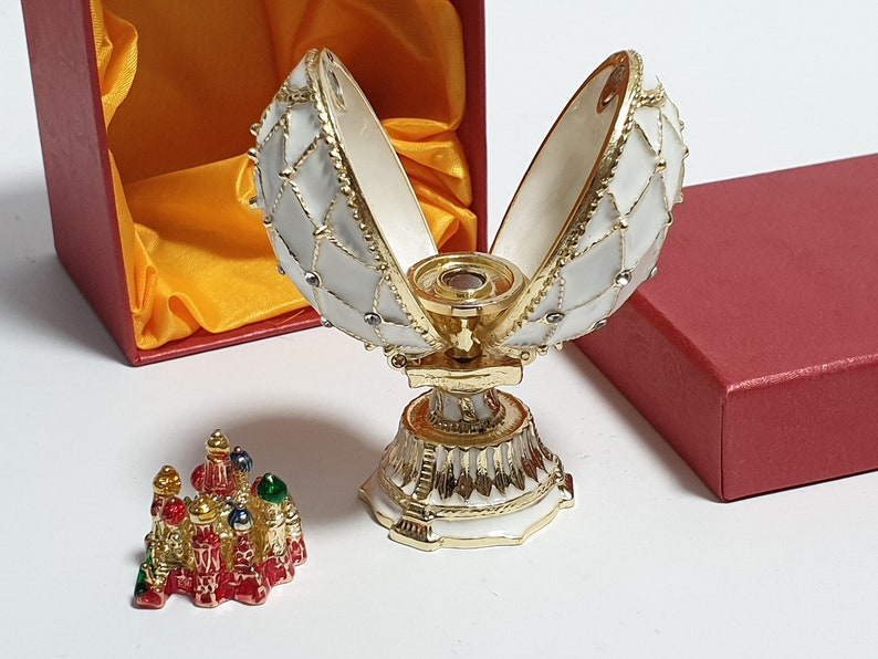 May include: A white and gold Faberg&eacute; egg with a red and gold miniature building inside. The egg is open and the building is visible. The egg is on a white stand with gold trim. The egg is in a red box with a yellow satin lining.