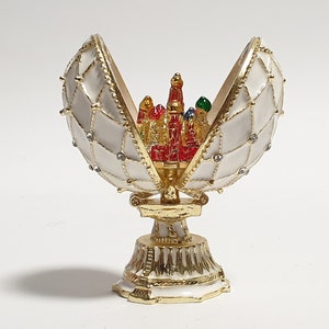 May include: A white and gold egg-shaped trinket box with a hinged lid, open to reveal a miniature red, gold, and green building inside. The box is decorated with gold trim and white enamel.