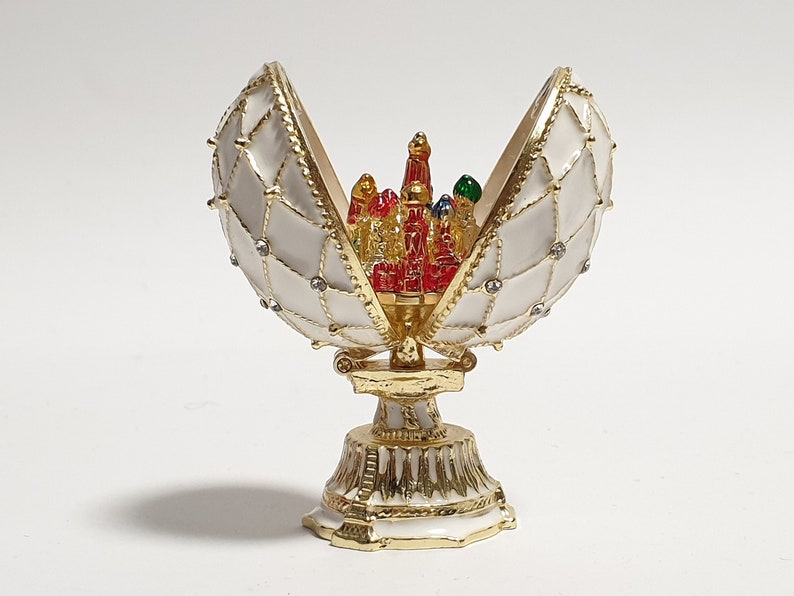 May include: A white and gold egg-shaped trinket box with a hinged lid, open to reveal a miniature red, gold, and green building inside. The box is decorated with gold trim and white enamel.