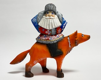 Op de afbeelding: Een handgeschilderd houten beeldje van een man in een rood, blauw en wit kostuum dat op een wolf rijdt. De man heeft een witte baard en een grijze hoed. De wolf is oranje en heeft zwarte ogen.
