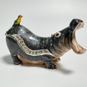 May include: A decorative hippo figurine with an open mouth, painted in shades of gray, black, and tan. A small yellow bird with a red beak sits on its back. The figurine is embellished with a row of clear rhinestones.