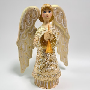 May include: A decorative angel figurine with a cream-colored dress and large wings. The angel is holding a gold trumpet. The wings and dress are adorned with gold and white decorative patterns. The angel has blonde hair and blue eyes.