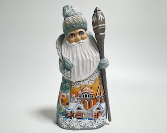 May include: A hand-carved wooden figurine of Santa Claus, dressed in a blue and white coat and hat, holding a staff. The coat features a detailed painting of a church and houses. The figurine has a long white beard and a smiling face.