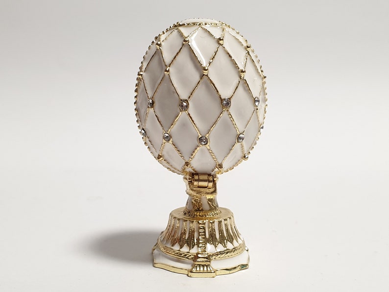 May include: A white porcelain egg with a gold lattice design and clear rhinestones. The egg is on a gold stand with a white base.