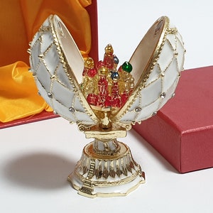 May include: A white and gold Faberg&eacute; egg with a red, green, and gold jeweled interior. The egg is open and the interior is visible. The egg is sitting on a gold stand.