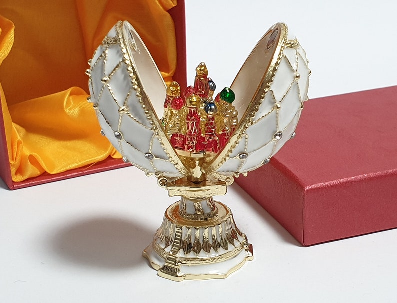 May include: A white and gold Faberg&eacute; egg with a red, green, and gold jeweled interior. The egg is open and the interior is visible. The egg is sitting on a gold stand.