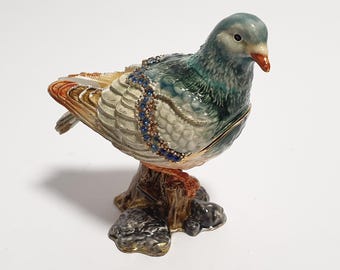 Op de afbeelding: Decoratief duifbeeldje met een blauwgroen lichaam, geplaatst op een boomstronkbasis. De vogel heeft gedetailleerde veren, een rode snavel en is versierd met blauwe edelsteenaccenten. Het beeldje is een verzamelobject.