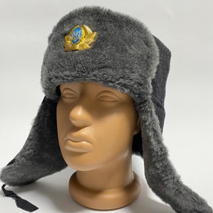 May include: A gray faux fur ushanka hat with ear flaps. The hat has a gold embroidered emblem with a blue trident on the front.
