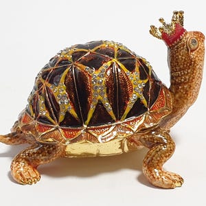 Könnte beinhalten: Dekorative Schildkrötenfigur mit Krone. Der Panzer der Schildkröte ist braun mit gelben und klaren Kristallakzenten. Die Krone ist goldfarben mit rotem Akzent. Die Figur ist aus Metall und etwa 10 cm lang.