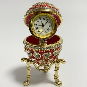 May include: A red and gold jeweled egg clock with a white face and Roman numeral hour markers. The egg is open and the clock face is visible. The egg is on a gold stand with three legs.