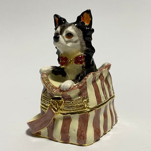 May include: A black and white chihuahua figurine wearing a red bow tie is peeking out of a striped, cream and brown, fabric bag. The bag is a trinket box with a gold clasp.