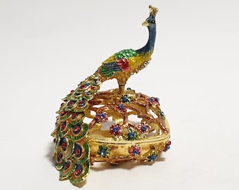 Puede incluir: Una caja de alhajas dorada con un diseño de pavo real. El pavo real es azul, verde y amarillo con detalles rojos. La caja está decorada con flores de colores y tiene una tapa con bisagras.
