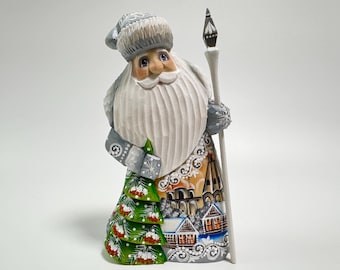 May include: A hand-carved wooden Santa Claus figurine. The figure has a white beard, blue eyes, and a gray hat. He holds a staff and is wearing a coat decorated with a Christmas tree and a winter village scene. The figurine is approximately 10 inches tall.
