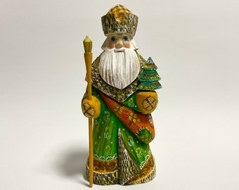 May include: A wooden figurine of a traditional Russian Santa Claus, or Ded Moroz, wearing a green and gold robe, a fur-trimmed hat, and holding a staff and a small green Christmas tree.