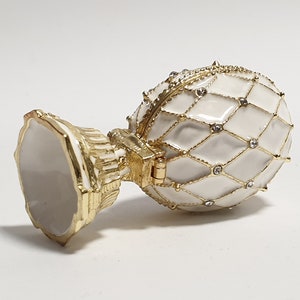 May include: A white porcelain egg-shaped trinket box with a gold-tone base and a diamond-shaped pattern. The lid is hinged and has a decorative design.