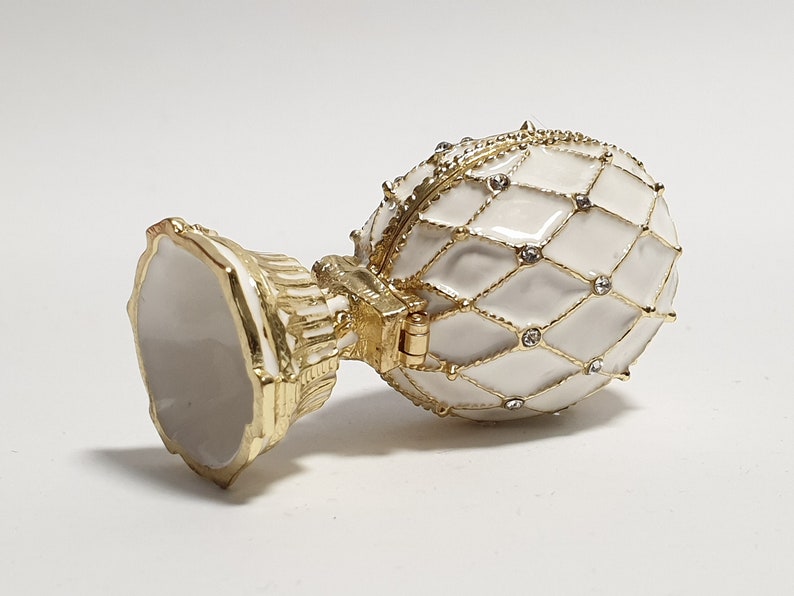May include: A white porcelain egg-shaped trinket box with a gold-tone base and a diamond-shaped pattern. The lid is hinged and has a decorative design.