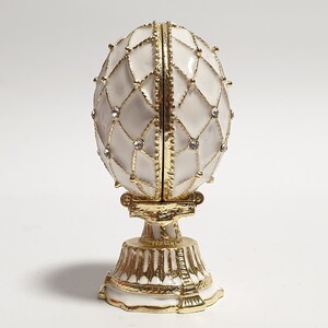 May include: A white porcelain egg with gold accents and clear rhinestones, on a gold stand. The egg is decorated with a geometric pattern.