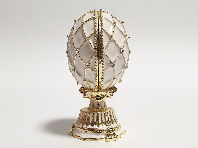 May include: A white porcelain egg with gold accents and clear rhinestones, on a gold stand. The egg is decorated with a geometric pattern.