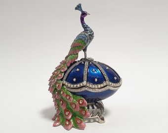 May include: A blue and green jeweled peacock figurine sits atop a blue egg-shaped music box with a pearl trim. The music box has a silver stand with three legs.