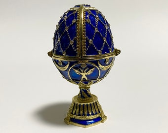 May include: A blue and gold Fabergé egg replica with a detailed design of a bird and intricate patterns. The egg is on a stand with a gold base and blue accents.