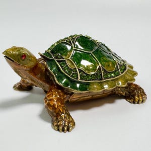 Può includere: Statuetta decorativa a forma di tartaruga con guscio verde tempestato di gioielli e corpo color bronzo. La tartaruga ha occhi di gemme rosse ed è appoggiata su una superficie bianca.