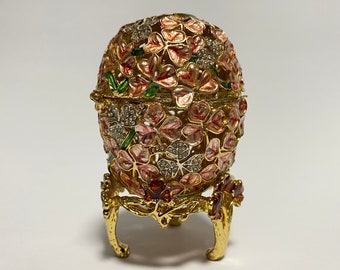 May include: A gold-tone decorative egg with a floral design. The egg is decorated with pink, white, and green enamel and has a stand with three legs.