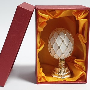 May include: A white and gold decorative egg with a diamond pattern and rhinestones, in a red box with a yellow satin lining.