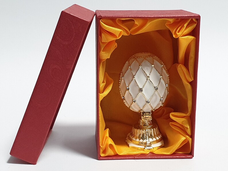 May include: A white and gold decorative egg with a diamond pattern and rhinestones, in a red box with a yellow satin lining.